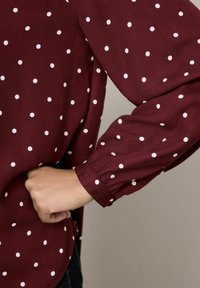 Bordowa bluzka w białe kropki, z długimi puff sleeves i zebranym mankietem. Gładka tekstura i lekko luźny krój w talii.