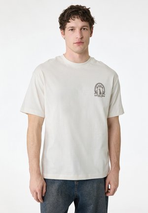 Jonge man met kort krullend donker haar, die een wit T-shirt met een kleine zwarte afbeelding en een blauwe spijkerbroek draagt, staand tegen een effen achtergrond.
