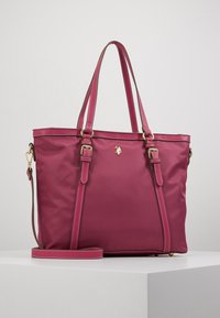 Różowa torebka w odcieniu malinowym z podwójnymi uchwytami u góry, regulowanym paskiem na ramię, złotymi klamrami oraz małym złotym emblematem na środkowej części przodu.