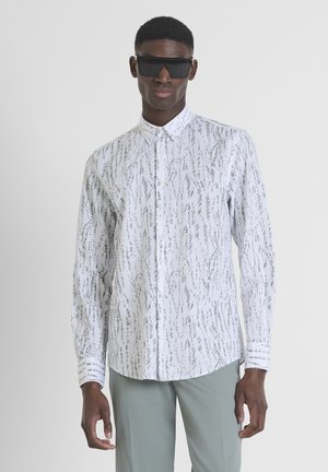 Camisa de manga larga blanca con un patrón abstracto en gris, con cuello abotonado y puños. Modelo que lleva gafas de sol negras, combinadas con pantalones verde claro.