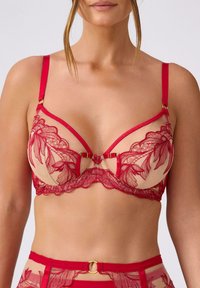 Czerwona przezroczysta koronkowa biustonosz z kwiatowymi wzorami, regulowanymi ramiączkami i akcentami z hardware'u w złotym odcieniu na cielistym tle.