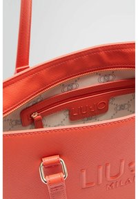 Borsa corallo arancione con esterno testurizzato, scomparti con zip e interno con marchio con un motivo ripetitivo. Accessori in tonalità argento.