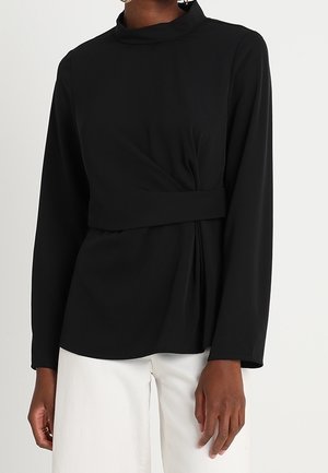 Kvinde iført en sort langærmet top med høj hals og detaljer med rynket talje, kombineret med hvide bukser med vide ben.