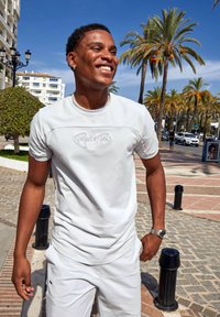 Camisa blanca de manga corta con logo bordado, combinada con pantalones cortos a juego. Tejido suave, diseño sencillo, ajuste relajado, al aire libre en un entorno soleado.