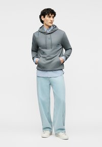 Νεαρός άνδρας που φοράει γκρι φούτερ με μπλε κέντημα, ανοιχτό μπλε ριγέ πουκάμισο, φαρδύ τζιν ανοιχτού μπλε χρώματος και λευκά αθλητικά, όρθιος με τα χέρια στις τσέπες.