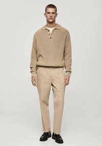 Beige gebreide pullover met een kraag en drie knopen, gecombineerd met lichtbeige broeken en zwarte schoenen, staand tegen een neutrale achtergrond.