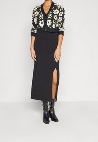 Schwarzer Maxirock mit seitlichem Schlitz, kombiniert mit einer taillierten, knopfartigen Strickjacke mit einem floralen Muster in Weiß und Grün auf einem dunklen Hintergrund.