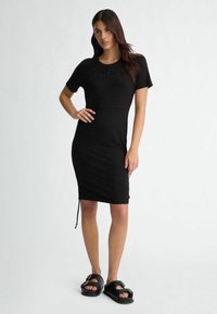 Black short-sleeve dress with a fitted silhouette, drawstring detail at the hem, and a textured logo on the chest. Paired with black sandals.