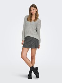 Mladi žena u sivoj pletenoj majici, tamnoj minici s uzorkom, crnim čarapama i crnim mokasinama stoji prekriženih nogu.