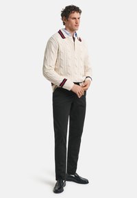 Cremfarbener Grobstrickpullover mit navy- und rotgestreiften Bündchen, kombiniert mit schwarzen Hosen und schwarzen Slippern, getragen von einem Mann mit lockigem Haar.