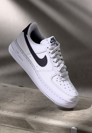 Hvid Nike Air Force 1 sneaker med sort swoosh logo, hvide snørebånd og perforeret tå, der vises svævende over en betonoverflade.