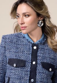 Veste en tweed bleu tiss�é avec un col en denim, deux poches foncées et des boutons en argent. Présente un détail d'ourlet effiloché le long du col.