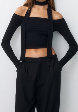 Femme portant un haut court noir à épaules dénudées avec des manches longues et un pantalon noir taille haute assorti, les mains dans les poches.