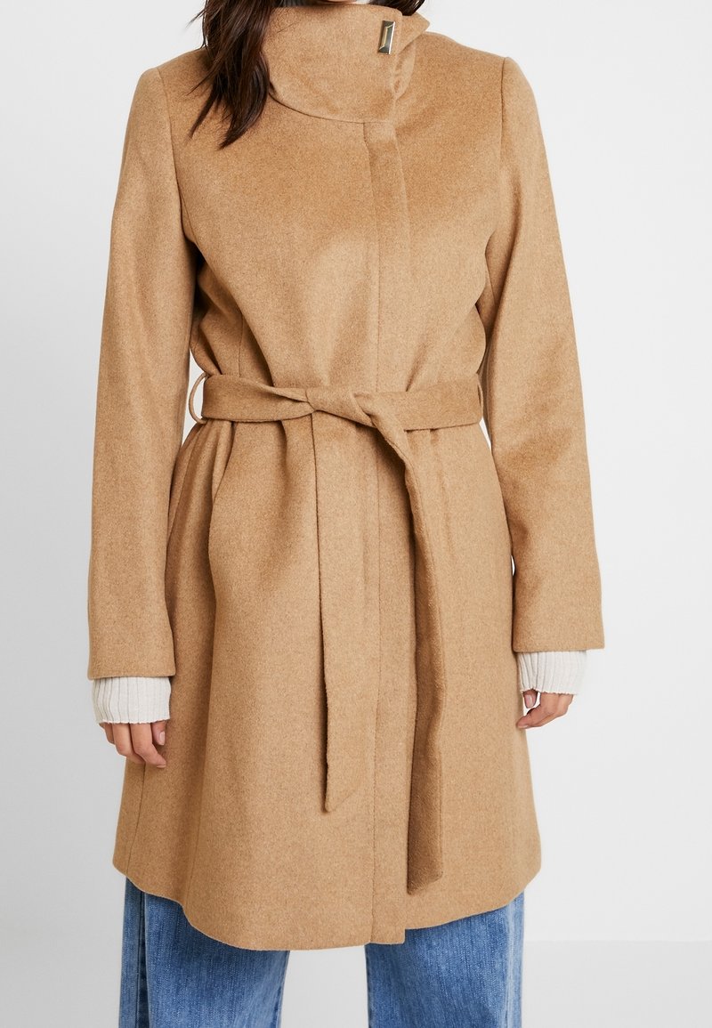 Femme portant un manteau en laine couleur camel avec un col haut et une ceinture nouée, par-dessus un jean bleu et des poignets blancs côtelés visibles aux manches.