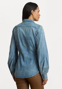 Ljusblå denimskjorta med långa ärmar, krage och en figursydd passform. Har sömnadsdetaljer och knappade ärmslut.