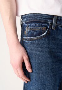 Σκούρο μπλε τζιν με κλασική εφαρμογή, με πορτοκαλί ραφές και μια μικρή ετικέτα "BOSS". Το χέρι resting στο πλάι της τσέπης.