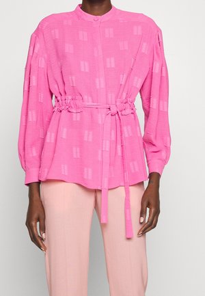 Femme portant un chemisier rose vif à manches longues avec une ceinture, associé à un pantalon rose clair, debout devant un fond uni.