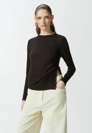 Maglia a maniche lunghe marrone scuro con scollatura a girocollo; presenta arricciature laterali e bottoni decorativi, abbinata a pantaloni in velluto a coste di colore chiaro.