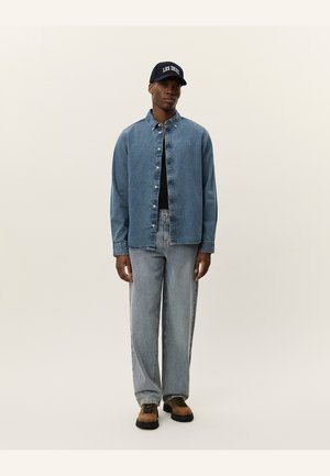 Denim skjorte i lyseblå med knaplukning, parret med lyseblå lige ben jeans, en mørk skjorte under, og en marineblå kasket med hvid tekst.