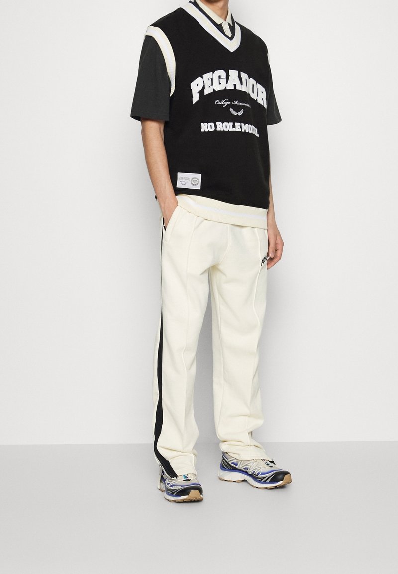 Personne portant une chemise noire à manches courtes, un pull sans manches noir et blanc avec du texte, un pantalon crème avec une bande noire et des chaussures de sport bleues.