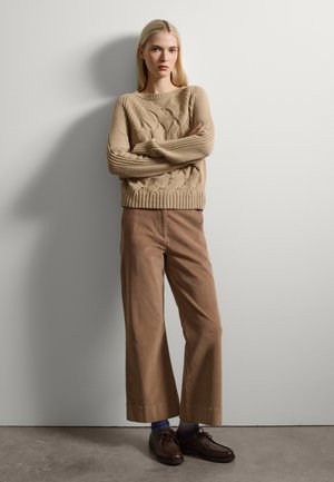 Beiger, kabelgestrickter Pullover mit langen Ärmeln; hellbraune, weite Cordhosen; dunkelbraune Schuhe und mehrfarbige Socken sichtbar.