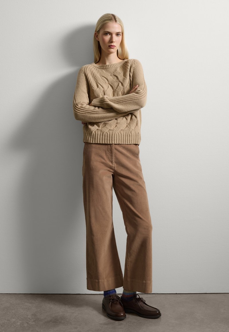 Suéter beige de punto con manga larga; pantalones de pana anchos de color marrón claro; zapatos marrón oscuro y calcetines multicolores visibles.