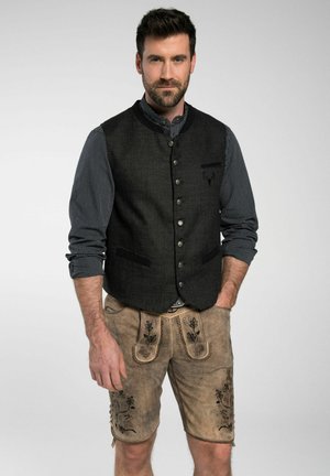 Schwarze Wollenweste mit Knopfverschluss, kombiniert mit hellbraunen Ledershorts mit floralen Stickereien und Taschen. Darunter ein gestreiftes Shirt.
