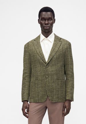 Man draagt een groene gestructureerde blazer over een wit overhemd met knopen en lichtbruine broek, staand tegen een eenvoudige lichte achtergrond.