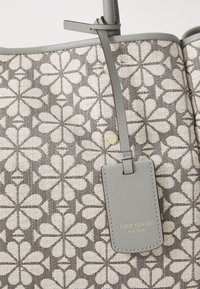 Siva torba s kremastim cvjetnim uzorkom, mekane tkanine i srebrna kožna oznaka na kojoj piše "kate spade NEW YORK" zlatnim slovima.