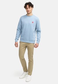 Felpa azzurro chiaro in misto cotone, con scollatura rotonda e piccolo logo rosso sul petto, abbinata a pantaloni beige e sneakers bianche.
