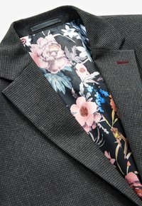 Grå tekstureret blazer med blomstermønsteret foer i nuancer af pink, blå og grøn. Har en struktureret krave og subtile røde detaljer.