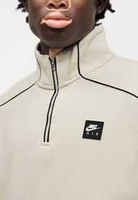 Μπεζ φούτερ με φερμουάρ μέχρι τη μέση, κατασκευασμένο από απαλό ύφασμα, με μαύρες λεπτομέρειες κατά μήκος των ραφών και τετράγωνο λογότυπο Nike Air στο στήθος.