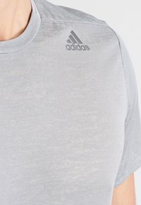 Camiseta deportiva de color gris claro hecha de tejido texturizado, con cuello redondo y un sutil logotipo tonal de Adidas en el hombro izquierdo.