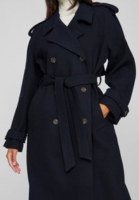 Γυναίκα που φοράει ένα σκούρο ναυτικό μπλε παλτό με διπλό κούμπωμα, ζώνη και μεγάλες παλτό, ποζάρει με το ένα χέρι στην τσέπη.