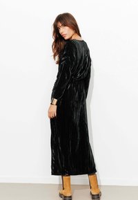 Robe longue en velours noir avec une finition texturée, manches longues et dos drapé. Associée à des bottines couleur sable et des accents en métal doré.