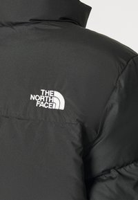 Czarna puchowa kurtka z wysokim kołnierzem i białym wyszywanym logo "The North Face" na górnej części pleców. Gładka tekstura, ocieplany fason.