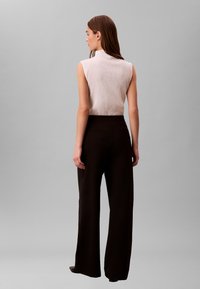 Frau mit langen braunen Haaren, die ein ärmelloses weißes Oberteil und hoch taillierte, schwarze Hosen mit weitem Bein trägt, steht mit dem Rücken zum Betrachter vor einem grauen Hintergrund.