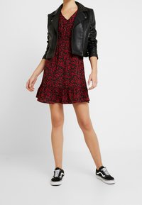 Giacca da biker nera in pelle sopra un vestito rosso con stampa floreale e orlo a campana. La modella indossa sneakers nere con dettagli bianchi.