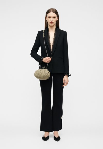 Γυναίκα με μαύρο σακάκι με δαντέλα στο τελείωμα, μαύρο καμπάνα παντελόνι με κουμπιά, μαύρα μυτερά παπούτσια, κρατώντας μπεζ τσάντα με ραβδώσεις και αλυσίδα λουράκι.
