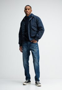 Marineblauer gepolsterter Mantel mit Fronttaschen, kombiniert mit blauen Jeans und schwarzen Sneakern. Der Mantel verfügt über einen Reißverschluss und Druckknöpfe.