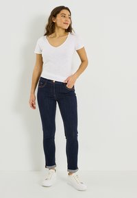 T-shirt blanc à manches courtes avec col en V, associé à un jean slim bleu marine, manches retroussées, et des baskets blanches à semelles texturées.