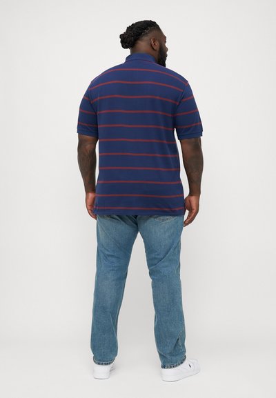 Marineblauwe poloshirt met rode horizontale strepen, korte mouwen en een kraag, gecombineerd met lichtblauwe jeans en witte sneakers.