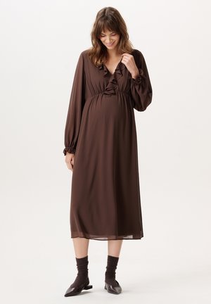 Vrouw in een bruine midi-jurk met lange mouwen en ruches, draagt donkere sokken en puntige schoenen, glimlacht en kijkt naar beneden.