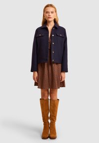 Giacca in misto lana navy con due tasche sul petto e bottoni dorati, indossata sopra un vestito marrone a fantasia e abbinata a stivali al ginocchio color sabbia.