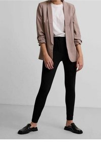 Světlý béžový blazer přes bílý tričko, zkombinovaný s černými legínami s vysokým pasem a černými mokasíny; elegantní design, střižený střih.