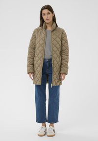 Khakifarbene steppjacke mit Stehkragen, goldenem Reißverschluss und langen Ärmeln, kombiniert mit einem gestreiften Hemd und hochgeschnittenen Jeans.