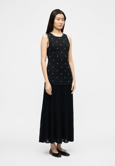 Νεαρή γυναίκα όρθια, φοράει μαύρο αμάνικο τοπ με μικρά λευκά σχέδια και μακριά μαύρη πλισέ φούστα με μαύρες φλατ.