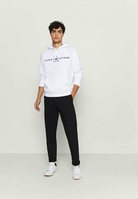 White hoodie with black text logo, drawstring hood, and ribbed cuffs, paired with black sweatpants and white sneakers. Minimalist design.