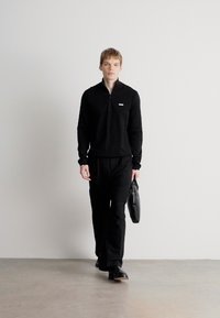 Sweater preto com fecho, adornado com um pequeno logotipo, combinado com calças pretas. O modelo segura uma mala preta e usa sapatos pretos sobre uma superfície lisa.