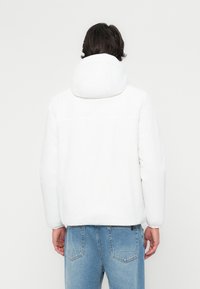 Hvid polstret jakke med hætte, der har en glat tekstur og et let design. Bæres med lyseblå denimjeans, der viser en afslappet pasform.